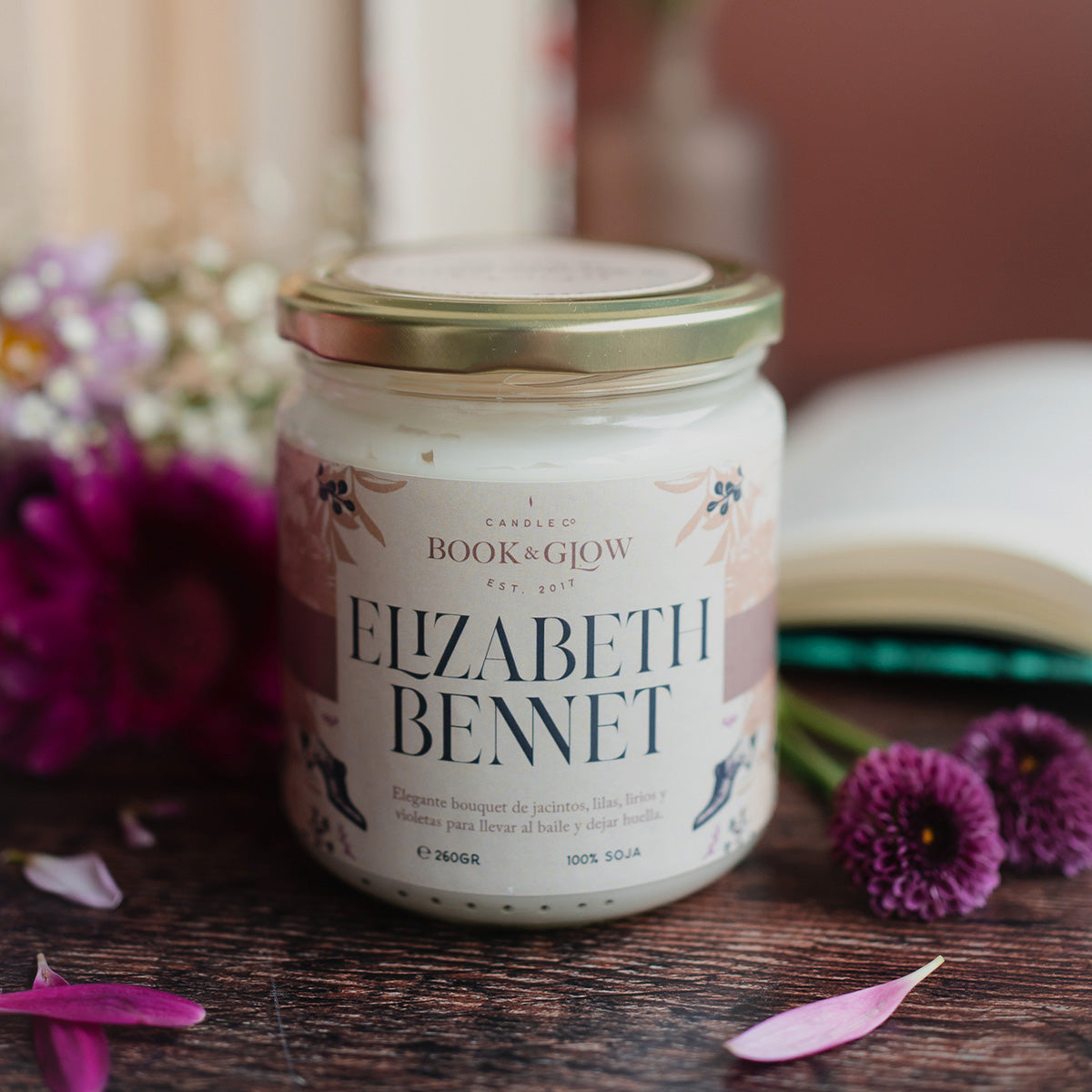 Vela de Book and Glow Elizabeth bennet, presentada en recipiente de cristal, etiqueta clara con título en color negro sobre mesa con flores y pétalos rosas.