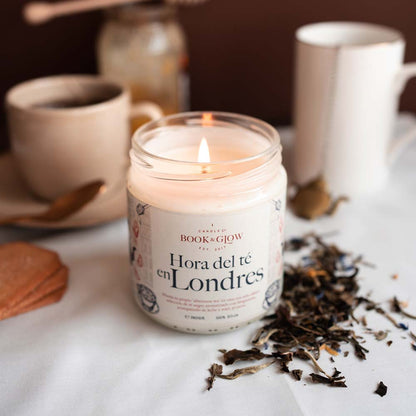Vela de Book and Glow Hora del té en Londres encendida, presentada en recipiente de cristal, etiqueta blanca con título en color negro sobre mesa con té negro.