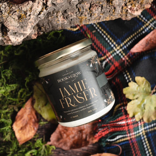 Vela de Book and Glow Jamie Fraser presentada en recipiente de cristal, etiqueta verde oscura con título en color amarilla sobre mesa con musgo y manta de tartán.