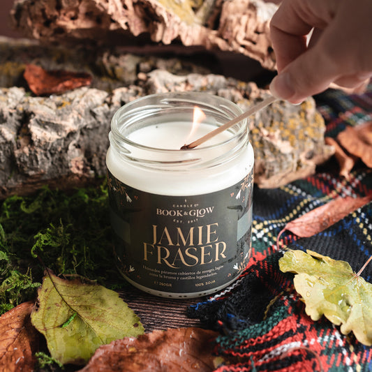 Vela de Book and Glow Jamie Fraser presentada en recipiente de cristal, etiqueta verde oscura con título en color amarilla sobre mesa con musgo y manta de tartán, siendo encendida con una cerilla.