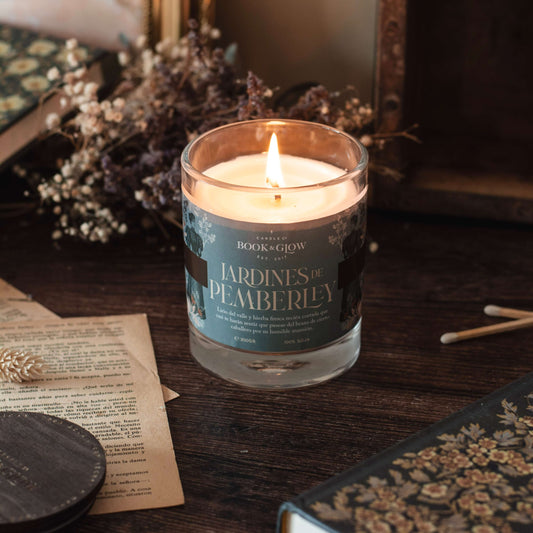 Vela de Book and Glow Jardines de Pemberley, presentada en recipiente de cristal abierto, etiqueta verde agua con título en color beige sobre mesa con flores y libros.