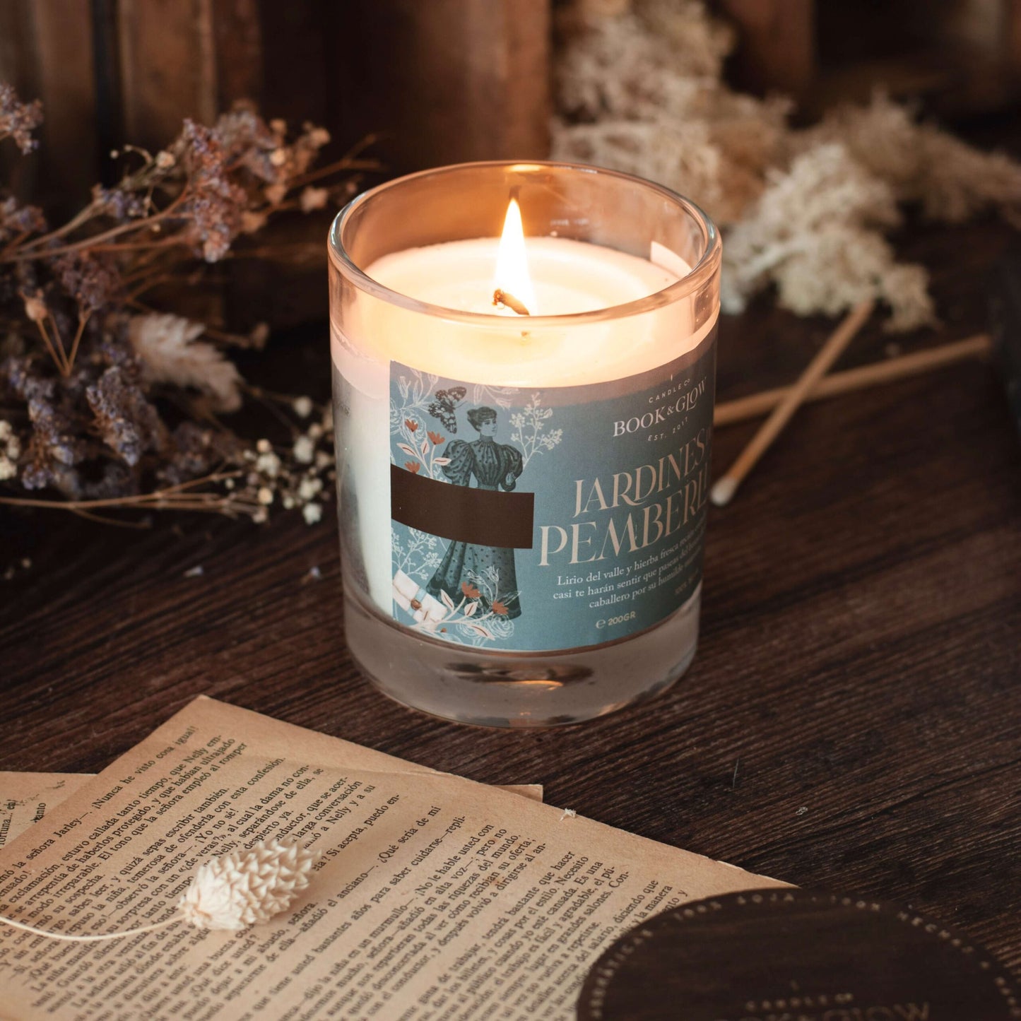 Vela de Book and Glow Jardines de Pemberley, presentada en recipiente de cristal, etiqueta verde agua con título en color beige sobre mesa con flores y libros de fondo.