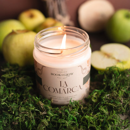 Vela de Book and Glow La Comarca encendida, presentada en recipiente de cristal, etiqueta clara con título en color verde oscuro sobre mesa con musgo y manzanas.