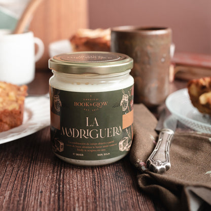 Vela de Book and Glow La Madriguera presentada en recipiente de cristal con etiqueta verde oscuro y título en beige sobre mesa de desayuno.