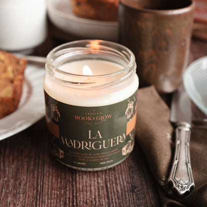 Vela de Book and Glow La Madriguera, encendida, presentada en recipiente de cristal con etiqueta verde oscuro y título en beige sobre mesa de desayuno.