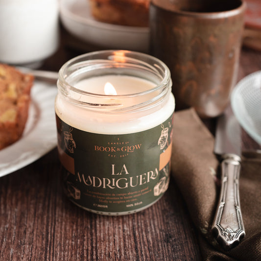 Vela de Book and Glow La Madriguera, encendida, presentada en recipiente de cristal con etiqueta verde oscuro y título en beige sobre mesa de desayuno.