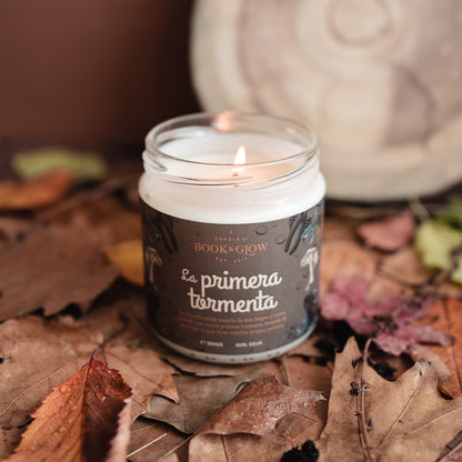 Vela de book and Glow La primera tormenta presentada en recipiente de cristal, etiqueta marrón con título en blanco sobre hojas secas.