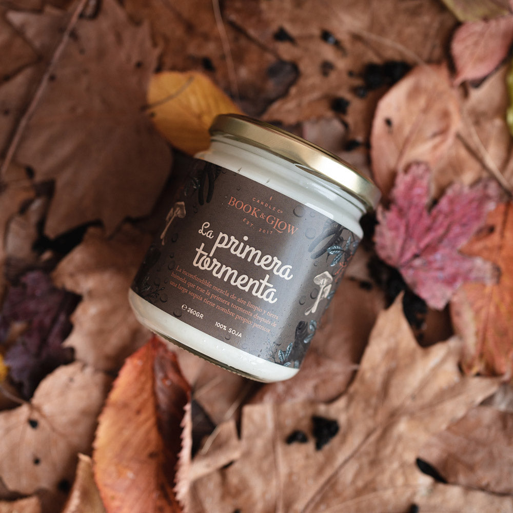 Vela de book and Glow La primera tormenta presentada en recipiente de cristal cerrado, etiqueta marrón con título en blanco sobre hojas secas.