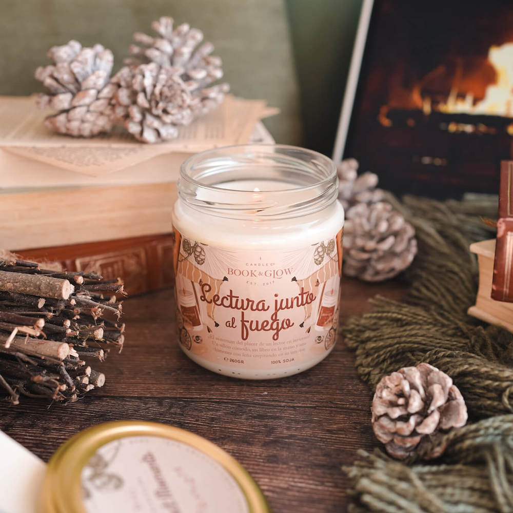 Vela de book and Glow Lectura junto al fuego presentada en recipiente de cristal, etiqueta en tonos anaranjados con título en naranja sobre mesa de madera.