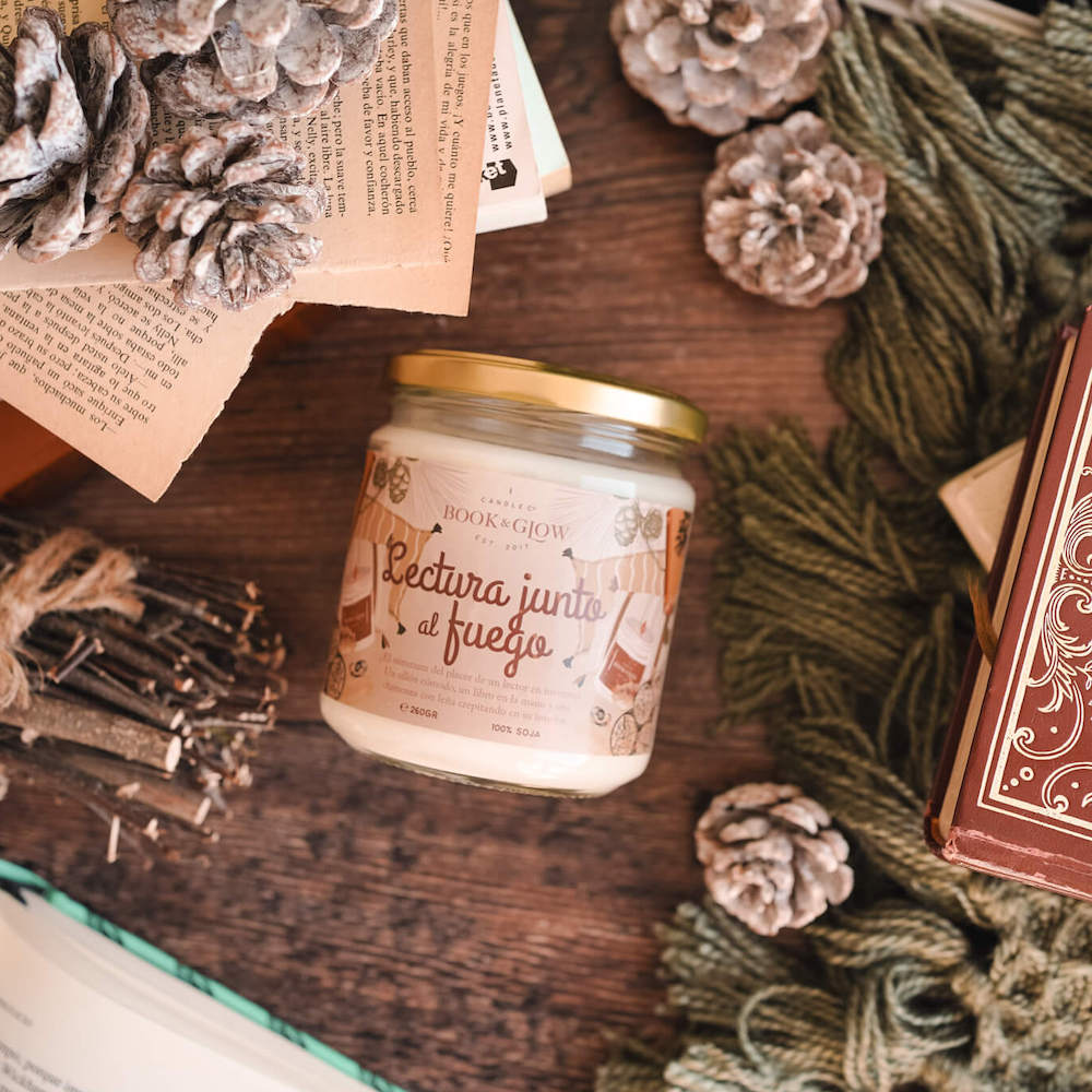 Vela de book and Glow Lectura junto al fuego presentada en recipiente de cristal cerrado, etiqueta en tonos anaranjados con título en naranja sobre mesa de madera.