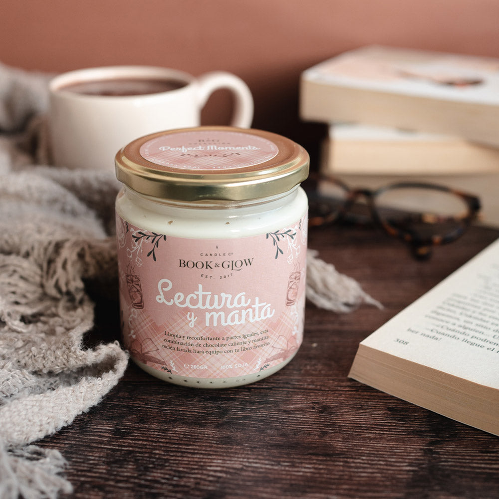 Vela de book and Glow Lectura y manta presentada en recipiente de cristal cerrado, etiqueta rosa con título en blanco sobre mesa de madera.