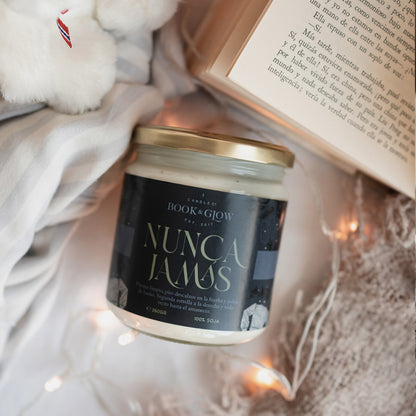 Vela de Book and Glow Nunca Jamás presentada en recipiente de cristal con etiqueta verde oscuro y título en beige sobre cama y junto a libro y manta.