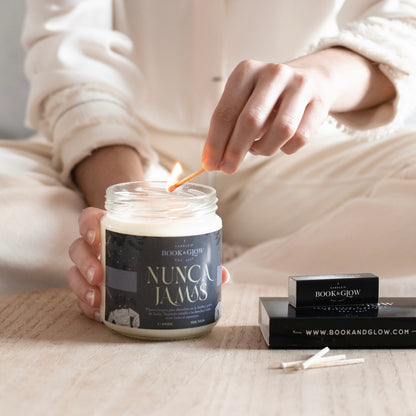 Vela de Book and Glow Nunca Jamás presentada en recipiente de cristal con etiqueta verde oscuro y título en beige, siendo encendida con una cerilla.