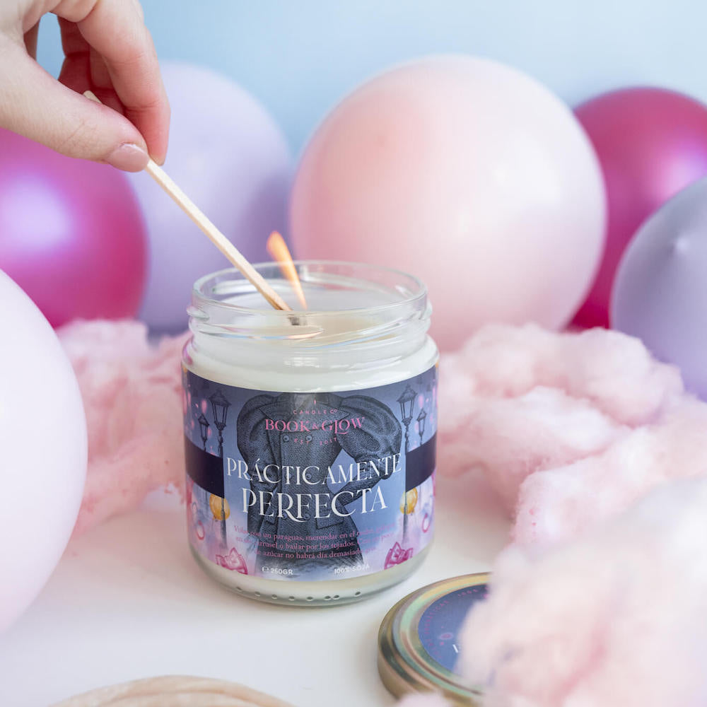 Vela de Book and Glow Prácticamente perfecta siendo encendida presentada en recipiente de cristal con etiqueta azul y título en blanco, sobre mesa llena de globos y algodón de azúcar.