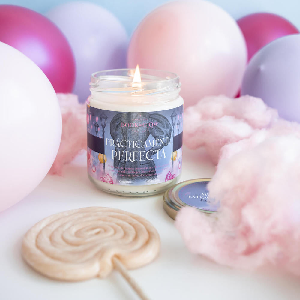 Vela de Book and Glow Prácticamente perfecta encendida presentada en recipiente de cristal con etiqueta azul y título en blanco, sobre mesa llena de globos y algodón de azúcar.