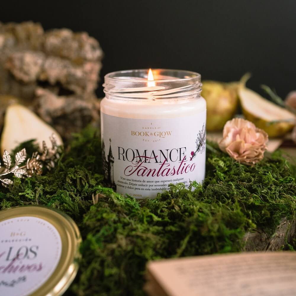 Vela de Book and Glow Romance Fantástico encendida, presentada en recipiente de cristal, etiqueta clara con título en color negro y rosa sobre mesa con musgo.