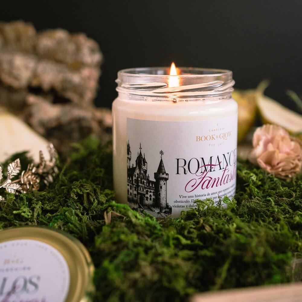 Lateral de la de Book and Glow Romance Fantástico encendida, presentada en recipiente de cristal, etiqueta clara con título en color negro y rosa e ilustración de castillo, sobre mesa con musgo.
