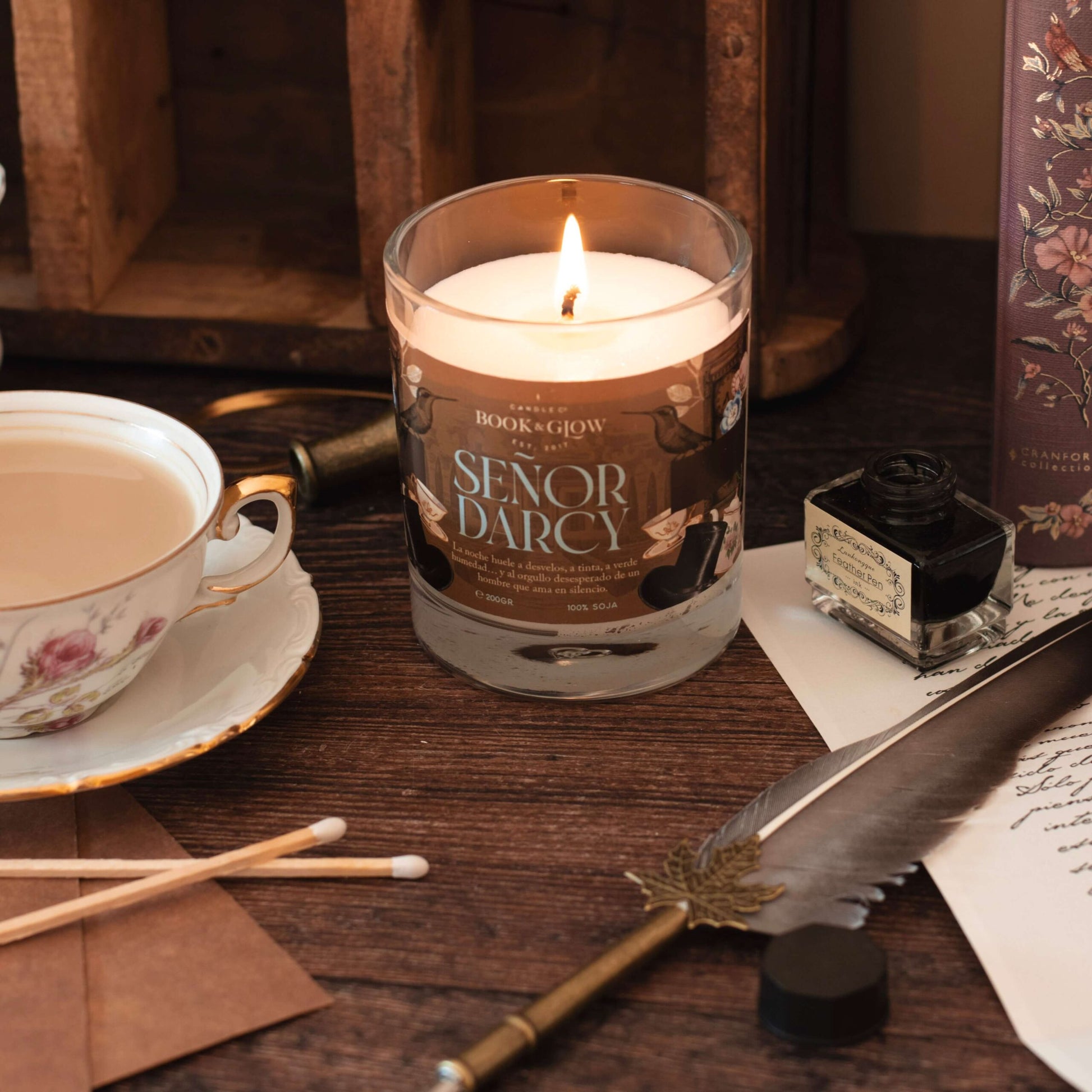 Vela Señor Darcy de Book and Glow encendida, presentada en recipiente de cristal junto a taza de té sobre escritorio de madera.